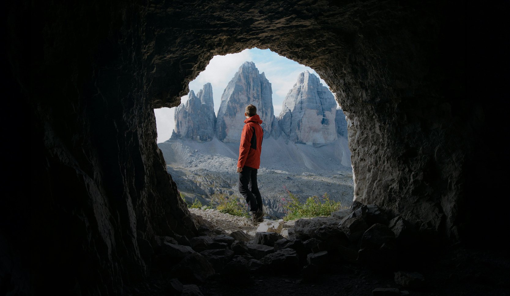 Man standing with his back turned to us at the center of the mouth of a cave.